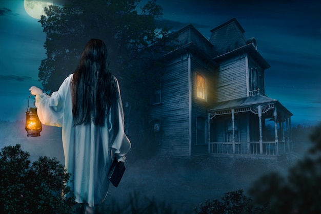 Female person in white shirt holds kerosene lamp and spellbook in hand against abondoned house, moonlit night, back view.