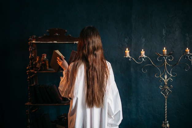 Female person in white shirt puts old spellbook on shelf, back view, candles. Dark magic, occultism and exorcism, witchcraft