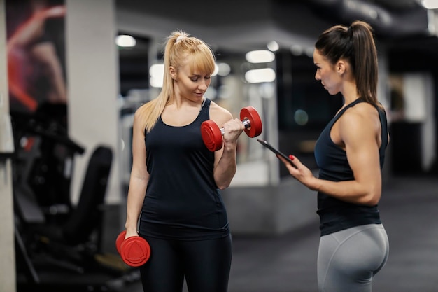 A female personal trainer is training fit sportswoman who is lifting dumbbells