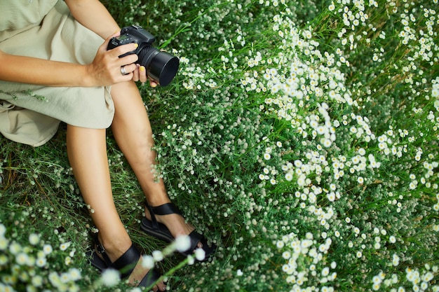 Female photographer sitting outdoors on flower field landscape holding a camera, unrecognizable woman hold digital camera in her hands. Travel nature photography, space for text, top view.