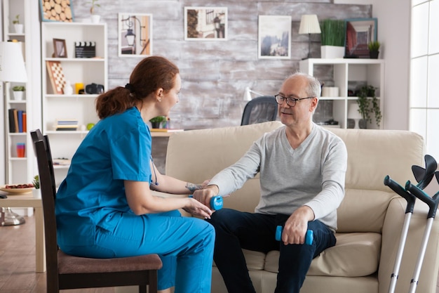 Female physiotherapist in assisting senior man lifting dumbbells in nursing home.