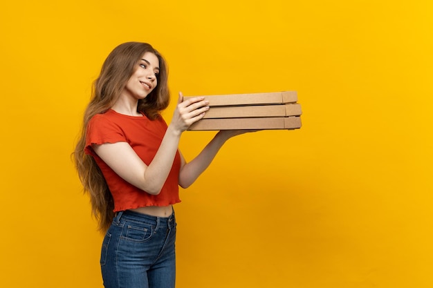 На этом фото изображена женщина-курьер пиццы, обрамленная ярким желтым фоном и несущая стопку коробок из-под пиццы.
