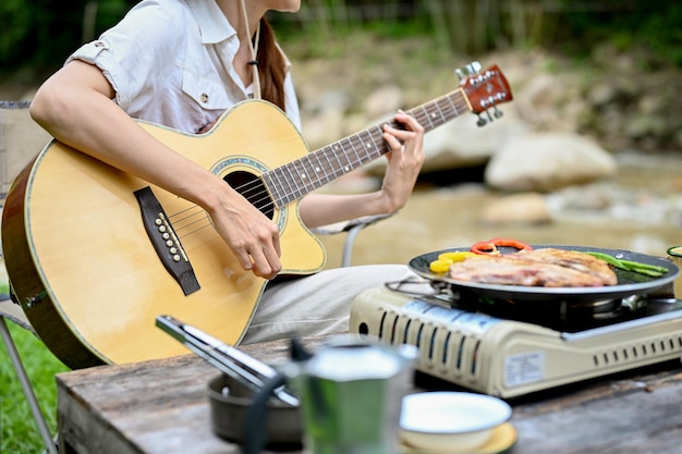 Female playing guitar while sitting near the river in the forest cropped image Camping concept