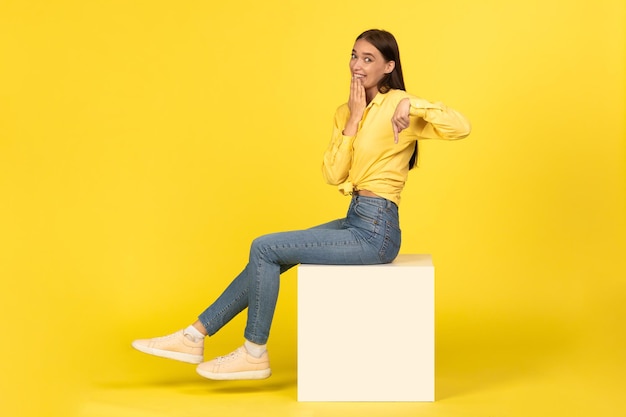 Female pointing finger down sitting on cube over yellow background