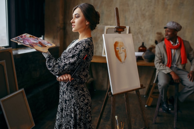 Female portrait painter holds palette and brush, male model in art studio. Male artist standing at his workplace, creative master in workshop