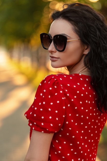 Female portrait of pretty young woman with fashion sunglasses in vintage summer red dress walk outdoors at sunset