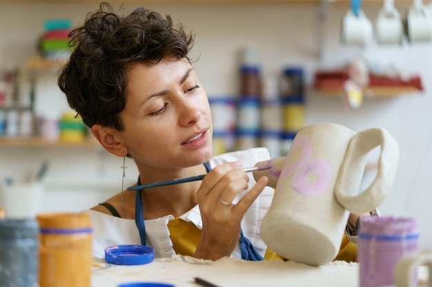 Female potter painting on clay jug in workshop working in pottery studio hobby small business