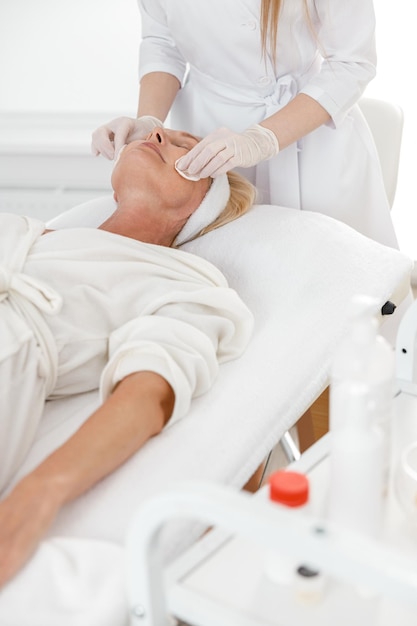 Female professional beautician applying facial skincare product on mature Caucasian woman