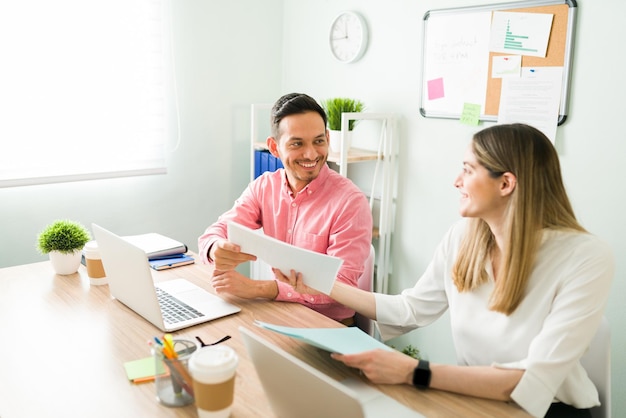 Female professional business partner smiling and giving a print work report to her male coworker while sitting at their office desk