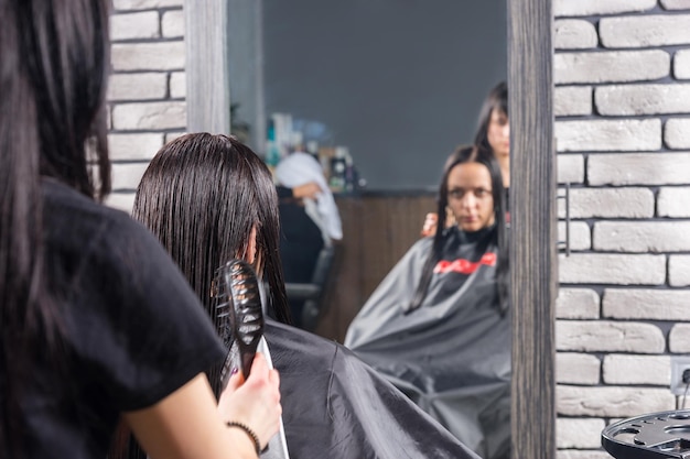Женский профессиональный стилист расчесывает мокрые волосы молодой клиентки брюнетки, пока она сидит в кресле в салоне красоты