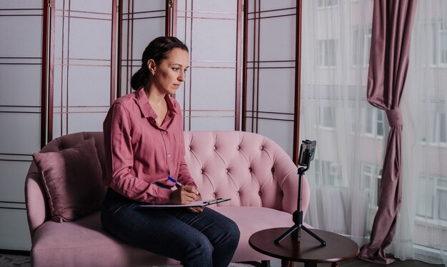 A female psychologist in business clothes sits on a pink sofa and conducts an online consultation using her phone