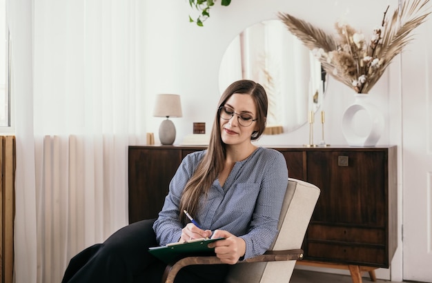 A female psychologist in glasses and a blue blouse makes a note on paper. The brunette is sitting in a chair in the room and taking notes. Psychological help.