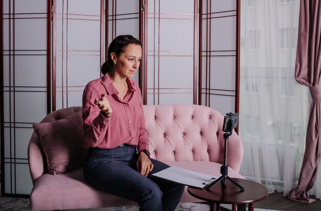 A female psychologist in a pink blouse is sitting on the couch and conducting an online consultation