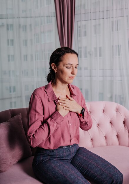 A female psychologist in a pink shirt is sitting on the sofa with her hands folded in a soothing gesture