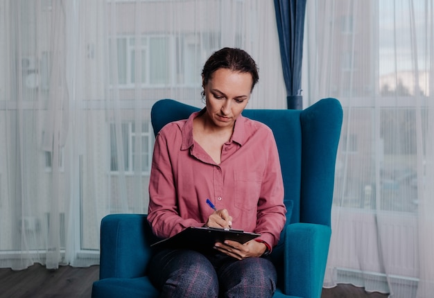 Женщина-психолог сидит на стуле в комнате и делает заметки