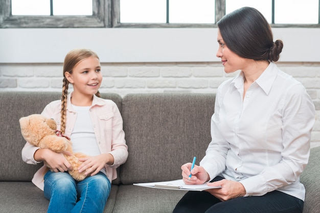 Female psychologist sitting with girl holding teddy bear taking note on clipboard