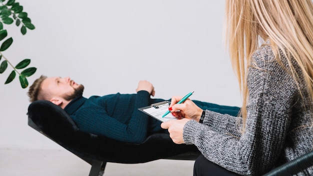 Female psychologist writing notes on clipboard in front of patient lying on couch in clinic