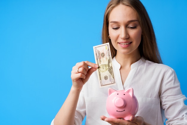 Женщина кладет деньги в розовую копилку на синем фоне