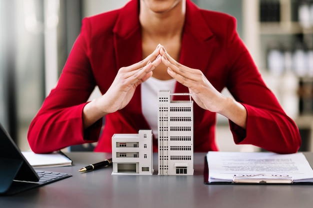 Female real estate agent is use hands to protect red roof for the concept of real estate investment about house trading purchase at desk in office xA