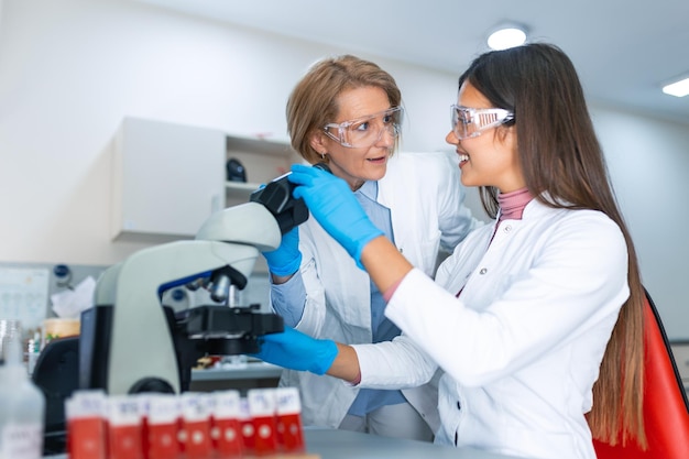 Female research scientist is analysing a sample on her microscope in a microbiology lab The lab is brightly lit with natural light Blurred glassware at side of frame provides copy space