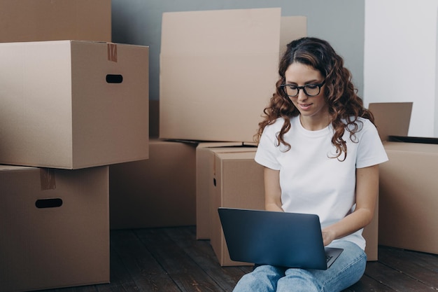 Female review moving company document on laptop online sitting\
on floor with boxes for relocation