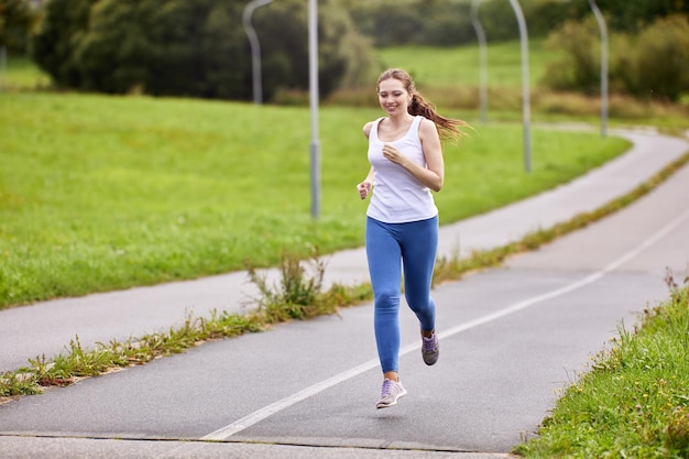 Female runner in white singlet and leggings makes morning jog along cycle track in city park in