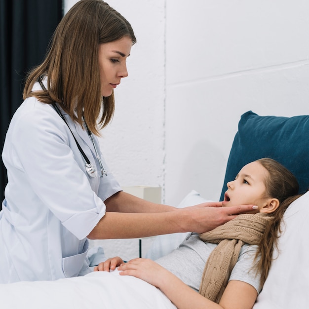 Foto il medico della femmina con lo stetoscopio intorno al suo collo che controlla la bocca di una ragazza malata che si trova sul letto