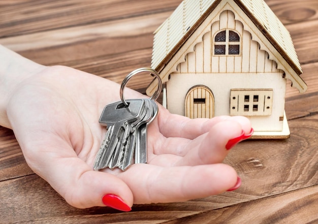 Female's hand holding keys for house and model of house on wooden background Mortgage and real estate concept