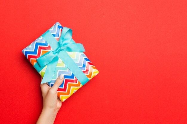 Female's hands holding striped gift box with colored ribbon on red. 