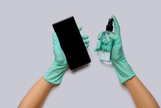 Female's hands in latex gloves wiping mobile phone with alcohol decontaminating agent top view over light grey background