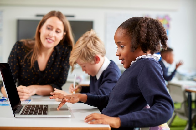 A female school teacher sitting at a desk helping a boy and girl using a laptop computer in primary school class side view