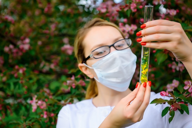 La scienziata nella mascherina medica con le provette in sue mani studia le proprietà delle piante in giardino botanico.