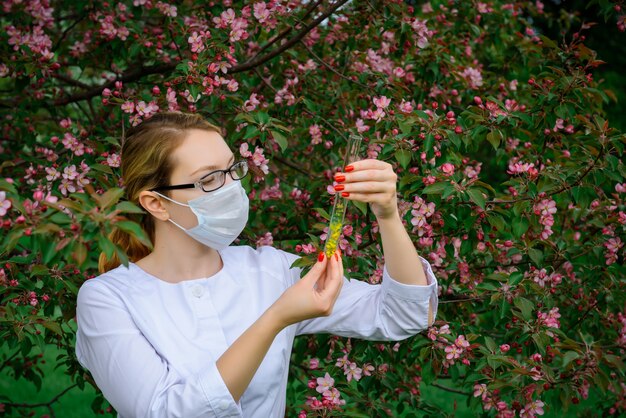 手に試験管を置いた医療用マスクの女性科学者は、植物園の植物の特性を研究しています。