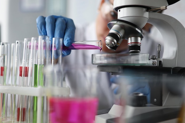 Female scientist pouring pink sample liquid on glass container under microscope in lab