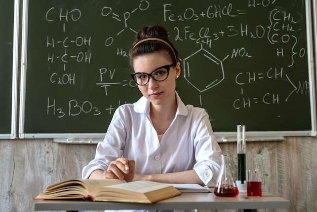 female scientist teacher with glasses in lab coat looking for chemical liquids in flasks in classroom againt blackboard