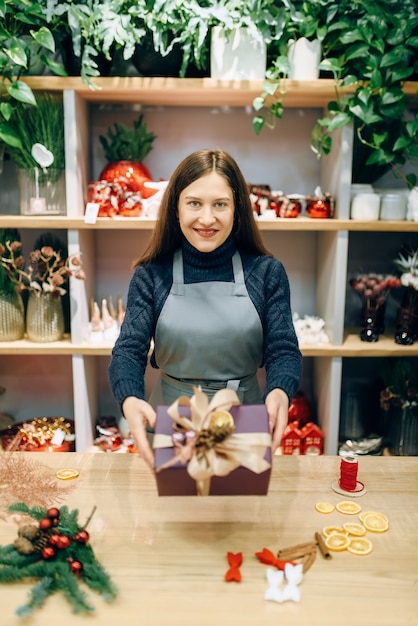 Female seller shows christmas gift box with handmade wrapping. Woman wraps present on the table, festive decor