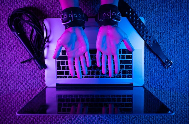 Photo the female sexy hands in bdsm handcuffs with laptop in neon light on dark background