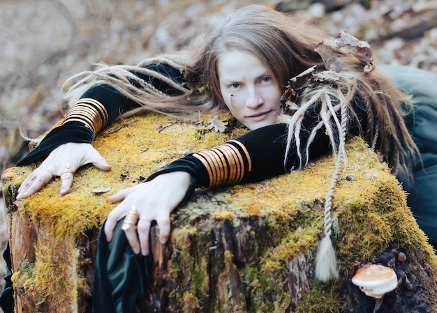 Photo female shaman in pristine forest nature laying on tree stump gold jewelry shaggy hair