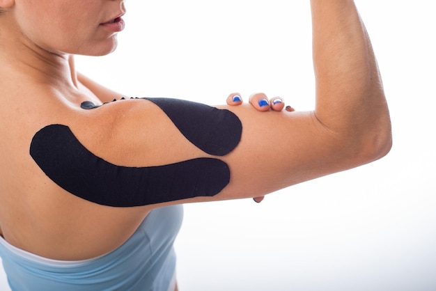 Foto spalla femminile con kineziotape su sfondo bianco medicina alternativa per infortuni sportivi delle articolazioni e dei legamenti giovane sportiva con un taping medico nero su una spalla dolorante da vicino