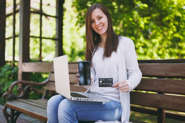 Female showing on camera credit card, mobile phone with blank empty screen to copy space. Woman sitting on bench working on modern laptop in street outdoors. Mobile Office. Freelance business concept.