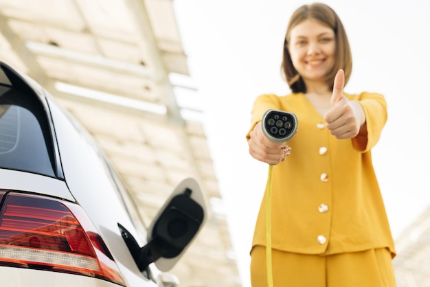 Female showing thumbs up holding power cable supply plugged at electric car charging station