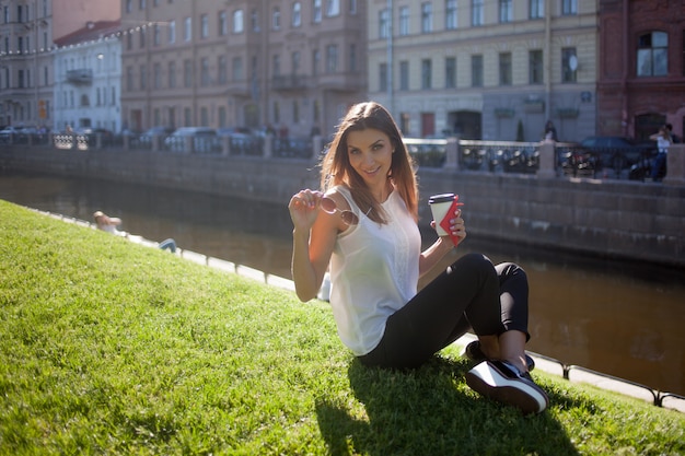 Женщина сидит на траве и пьет кофе в картонной чашке