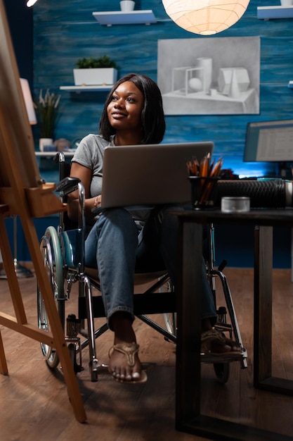 Female sketch artist wheelchair user using laptop computer to\
take drawing classes remotely at home made studio. african american\
looking for inspiration online to create artwork.