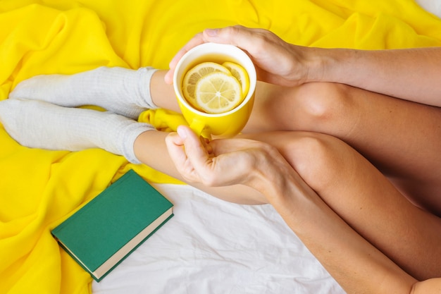 Female slender legs on a yellow plaid and a white sheet. In the hands of a yellow cup with tea and lemon. 