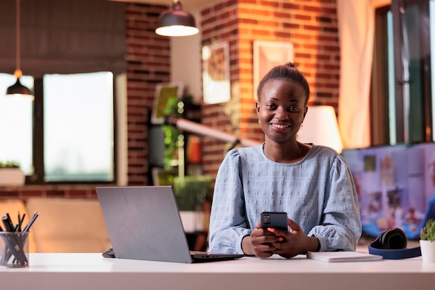 Female smiling remote worker using mobile phone and sitting at home office workplace. Successful african american SMM manager using smartphone and laptop and looking at camera