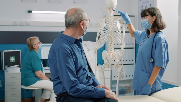 Female specialist pointing at spinal cord on human skeleton, explaining bones anatomy system to old patient at consultation. Assistant talking about osteopathy diagnosis, orthopedic spine.