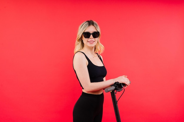 Female in sport clothes stands on electric scooter with sunglasses on red and white background