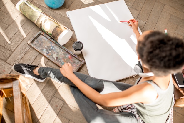 Female student drawing on the white canvas sitting on the floor. View from above