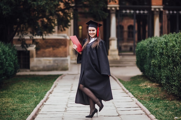 Female student graduate is standing in university hall in mantle, smiling and looking at the camera.