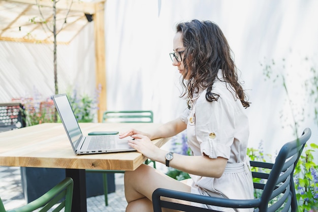 A female student sits on the summer terrace of a cafe and works at a laptop Female freelancer works remotely online while sitting in a summer cafe Remote work